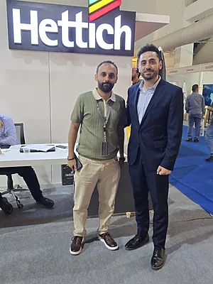 Two men, one in casual clothes and the other in a suit, posing under a Hettich sign at an event.