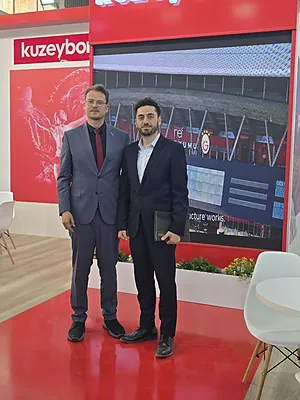 Two men in suits standing on a red floor in front of a digital display and red wall with white text.