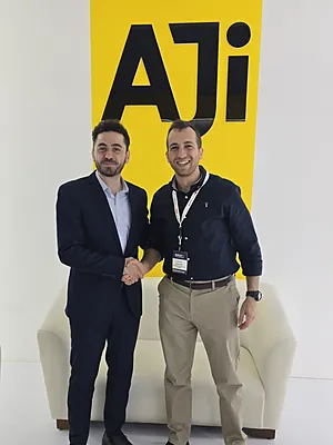 Two men shaking hands in front of a yellow wall with large black letters 'AJi'.