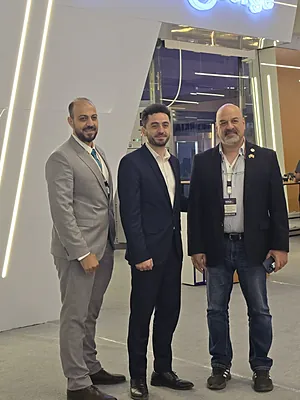 Three men in business attire standing in a modern office or conference space with a bright light strip on the wall.