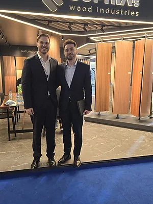 Two men in suits standing and posing in front of wood panel displays at a trade show booth.