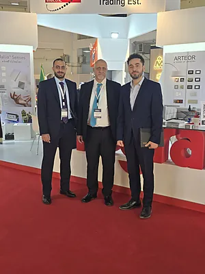 Three men in suits standing on red carpet at a trade show booth with promotional displays behind them.