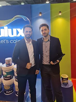 Two men in suits standing beside stacked Dulux paint cans at a colorful display booth.