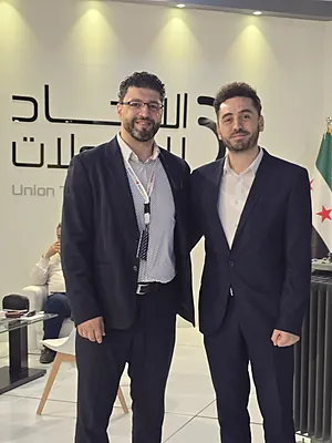 Two men in suits standing side by side in an office with a flag and Arabic text on the wall.