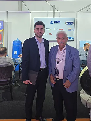 Two men standing and posing for a photo inside an exhibition booth with logos on a white wall behind them.