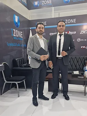 Two men in business attire standing in front of an IZONE Solutions booth with chairs and a table.