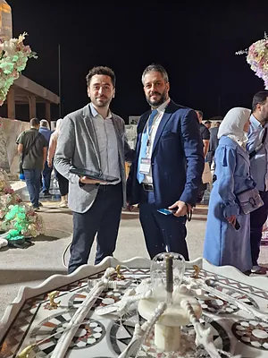 Two men in suits posing outdoors at night near a decorative table with floral arrangements and people in the background.