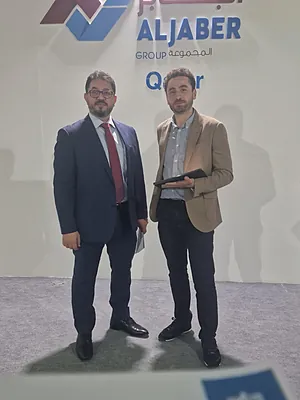 Two men in business attire posing in front of a wall with Aljaber Group Qatar logo.