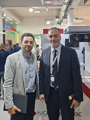 Two men standing indoors at a trade show or expo, both wearing business attire and smiling.
