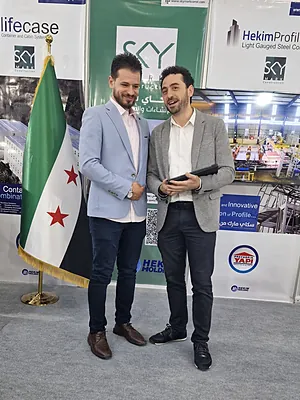 Two men in suits smiling and conversing in a trade show booth with a Syrian flag beside them.