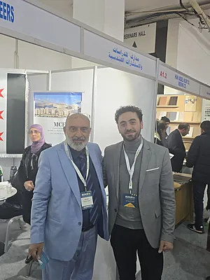 Two men with name badges standing and smiling in front of a booth at an indoor event.