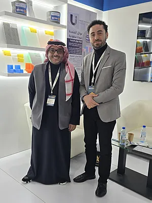 Two men standing indoors at a trade booth, one in traditional attire and the other in a gray blazer.