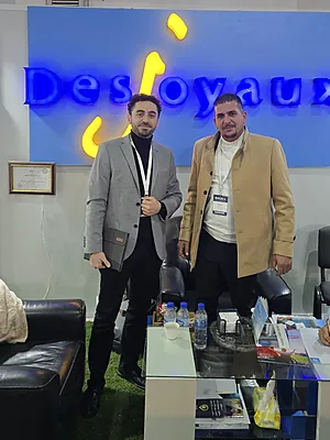 Two men standing inside an office with a blue wall sign and a glass table with water bottles.