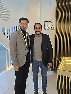 Two men wearing blazers stand side by side indoors near a Brand Aid sign and modern decor.