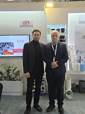 Two men in suits standing at a trade show booth with pipes and product displays behind them.