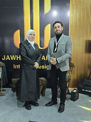Man in gray blazer and woman in black coat with white headscarf stand before gold and black Jawhar sign.