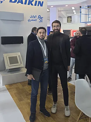 Two men standing and posing inside a Daikin booth at an indoor event.