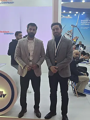 Two men in gray blazers standing at an exhibition booth with people and displays in the background.