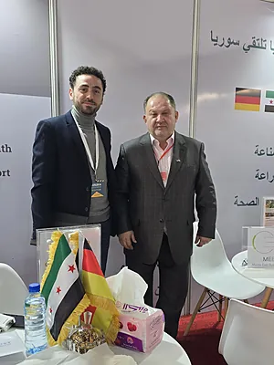 Two men in suits standing at a booth with Syrian and German flags and promotional materials on a table.