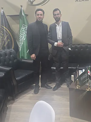 Two men in suits standing in a room with black leather sofas and Saudi Arabian flags.