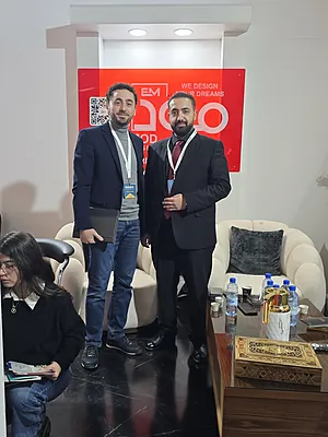Two men in business attire standing in front of a red sign and white chairs in an office setting.