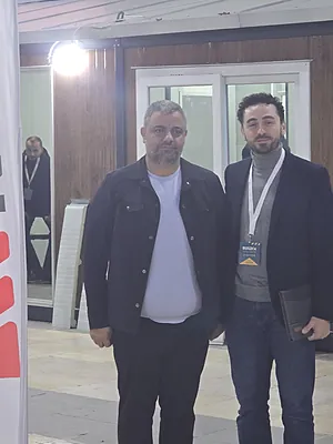 Two men standing indoors, one in a black jacket and the other in a blazer with a lanyard badge.