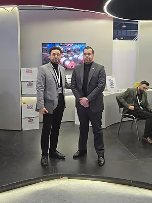 Two men in business attire standing in front of a booth with stacked boxes and a screen displaying barrels.