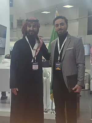 Two men standing side by side indoors, one in traditional Middle Eastern attire and one in a blazer with lanyards.