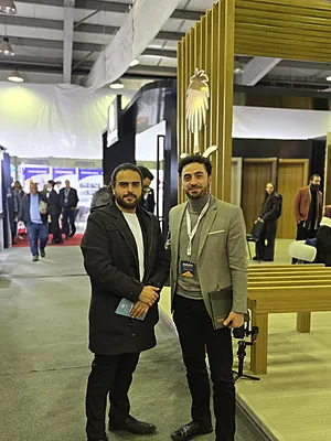 Two men standing inside a modern exhibition hall with wooden decor and people in the background.