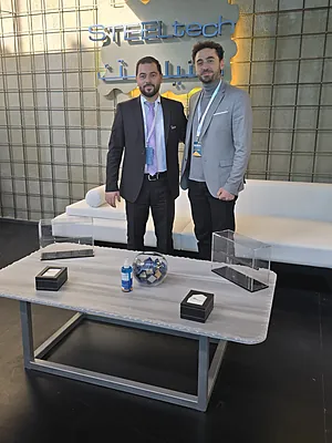 Two men in business attire stand behind a table with promotional items in front of a STEELtech sign.
