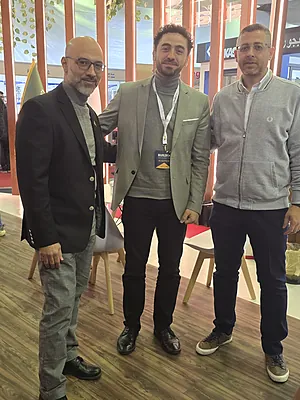 Three men standing indoors on wood floor, two wear blazers and one a gray zip-up jacket.