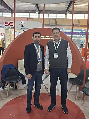 Two men standing inside a modern indoor setting with chairs and a curved orange backdrop behind them.