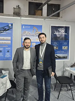 Two men in business casual clothing standing in front of construction-themed posters at an event booth.