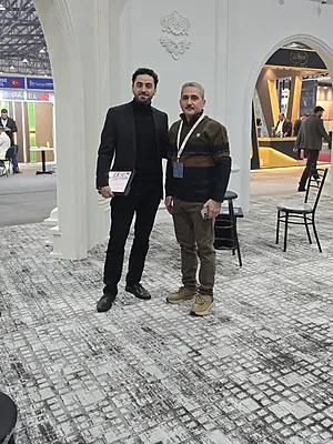 Two men standing indoors on a patterned carpet near white pillars and chairs in a convention or exhibit space.
