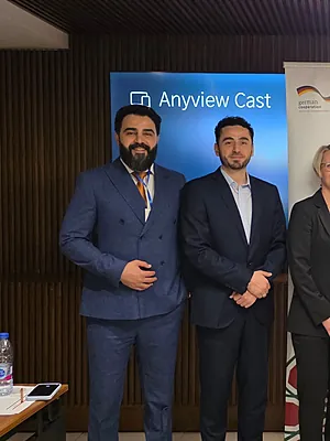 Two men in suits standing indoors in front of a screen displaying 'Anyview Cast'.